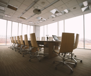 An empty, modern boardroom with a long wooden table and chairs, large windows, and a laptop on the table.